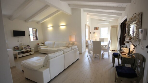 Uma sala de estar espaçosa com um sofá branco, uma área de jantar com mesa e cadeiras, e uma televisão fixada na parede.