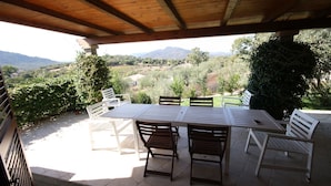 Um pátio externo com mesa e cadeiras de madeira, rodeado por vegetação e vista para o campo.