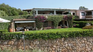 Una casa de dos pisos con un muro de piedra al frente, un espacio cubierto para sentarse al aire libre y un jardín bien cuidado con plantas en flor.
