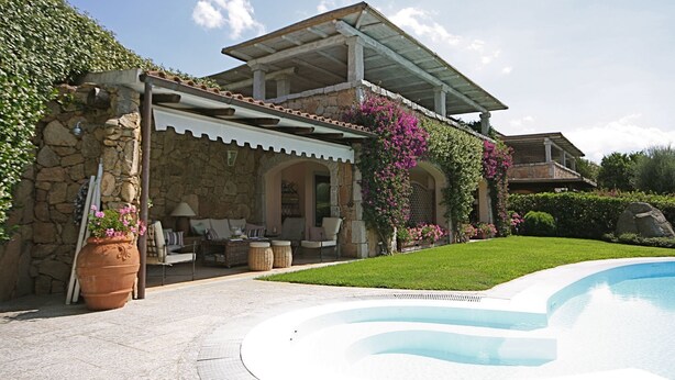 Una casa de piedra con un porche cubierto, piscina y abundante vegetación.