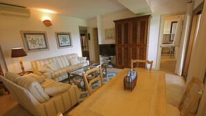 Una sala de estar con sofá, mesa de centro y mesa de comedor. Se ve un televisor y armarios de madera al fondo.