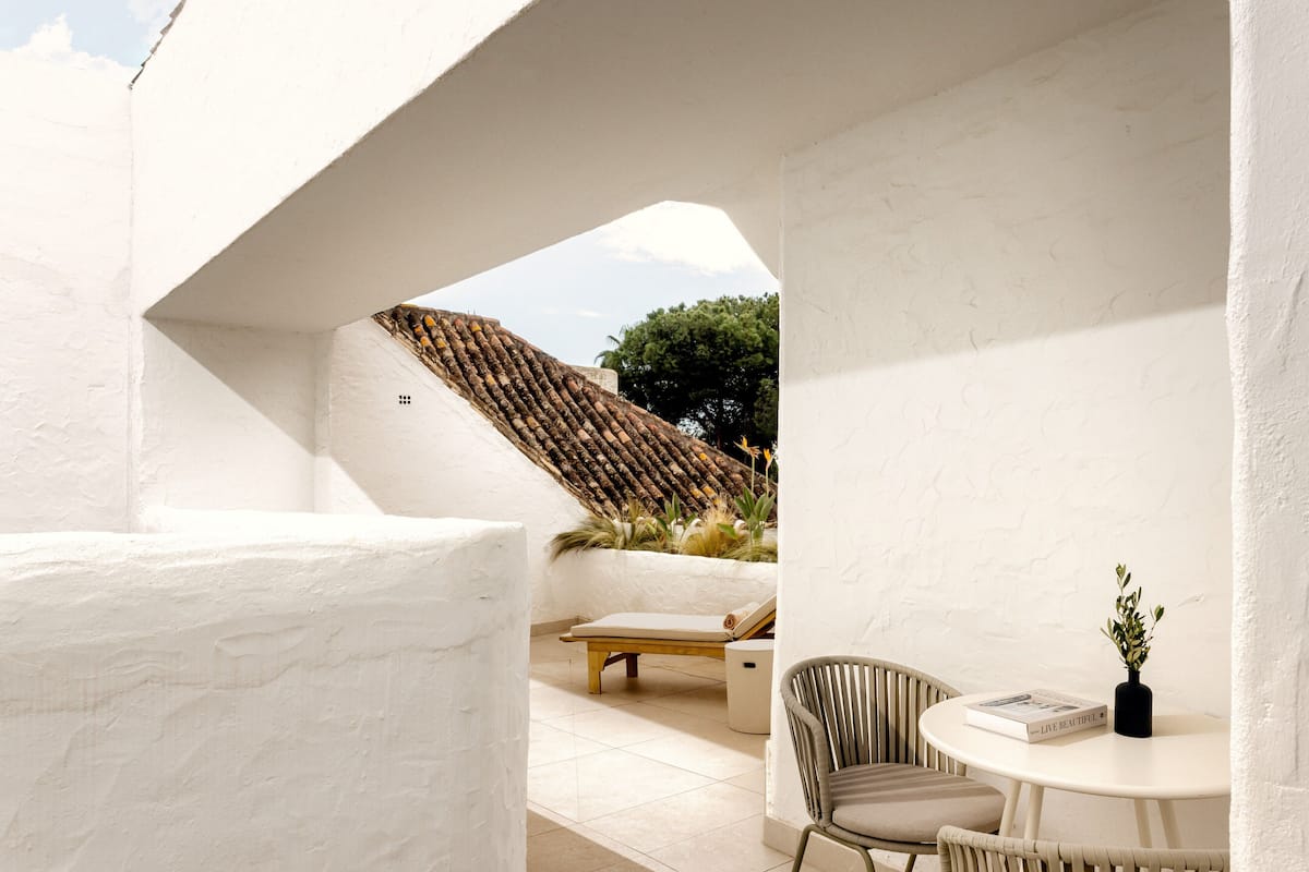 A modern outdoor seating area with a white wall, a wooden bench, and a round table with a book and a vase.