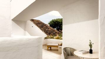 A modern outdoor seating area with a white wall, a wooden bench, and a round table with a book and a vase.