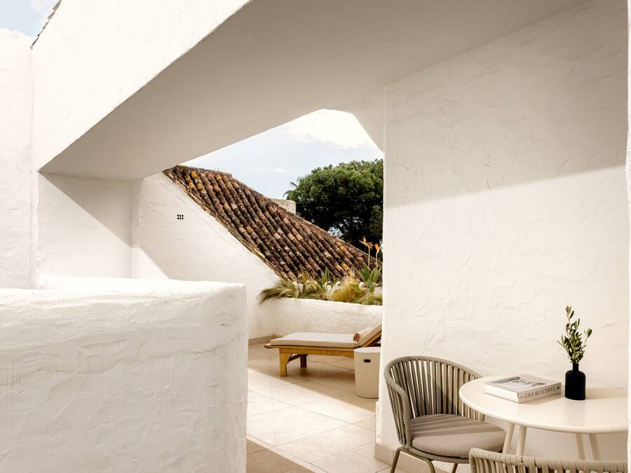 A modern outdoor seating area with a white wall, a wooden bench, and a round table with a book and a vase.