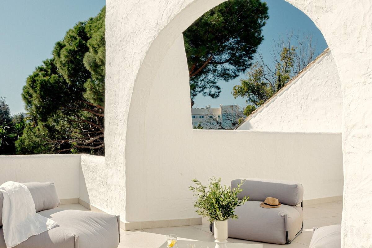 An outdoor seating area with a white textured wall, a sofa, a small table with drinks, and a potted plant.