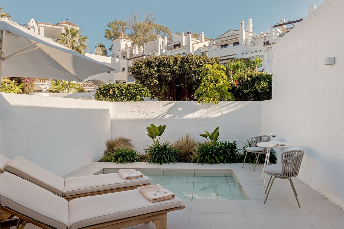 A rooftop pool area with lounge chairs and a table, surrounded by greenery and white walls.