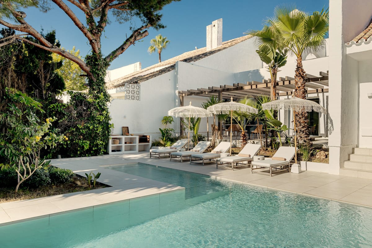 A pool area with lounge chairs and umbrellas, surrounded by greenery and a white building.