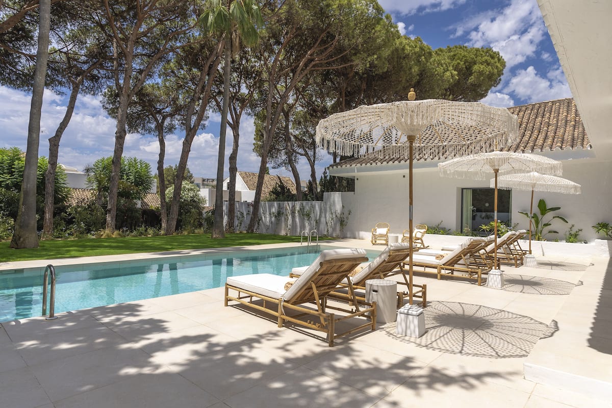 A poolside area with lounge chairs and umbrellas, surrounded by trees and a building with a tiled roof.