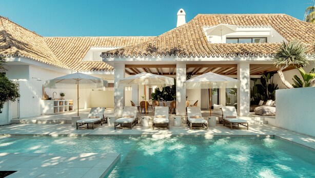Área da piscina com cadeiras de sol e guarda-sóis, telhado ladrilhado e fachada de edifício branca.