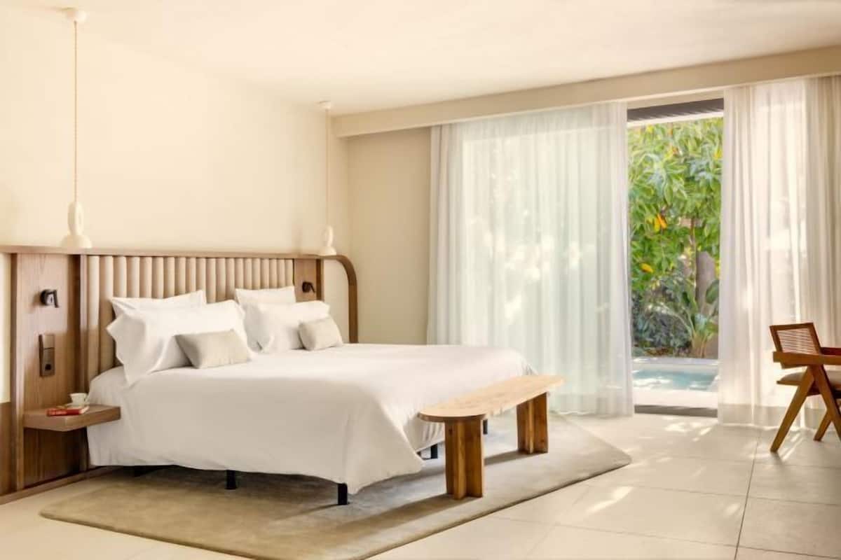 A bedroom with a bed, bedside table, bench, and a view of a pool and greenery.
