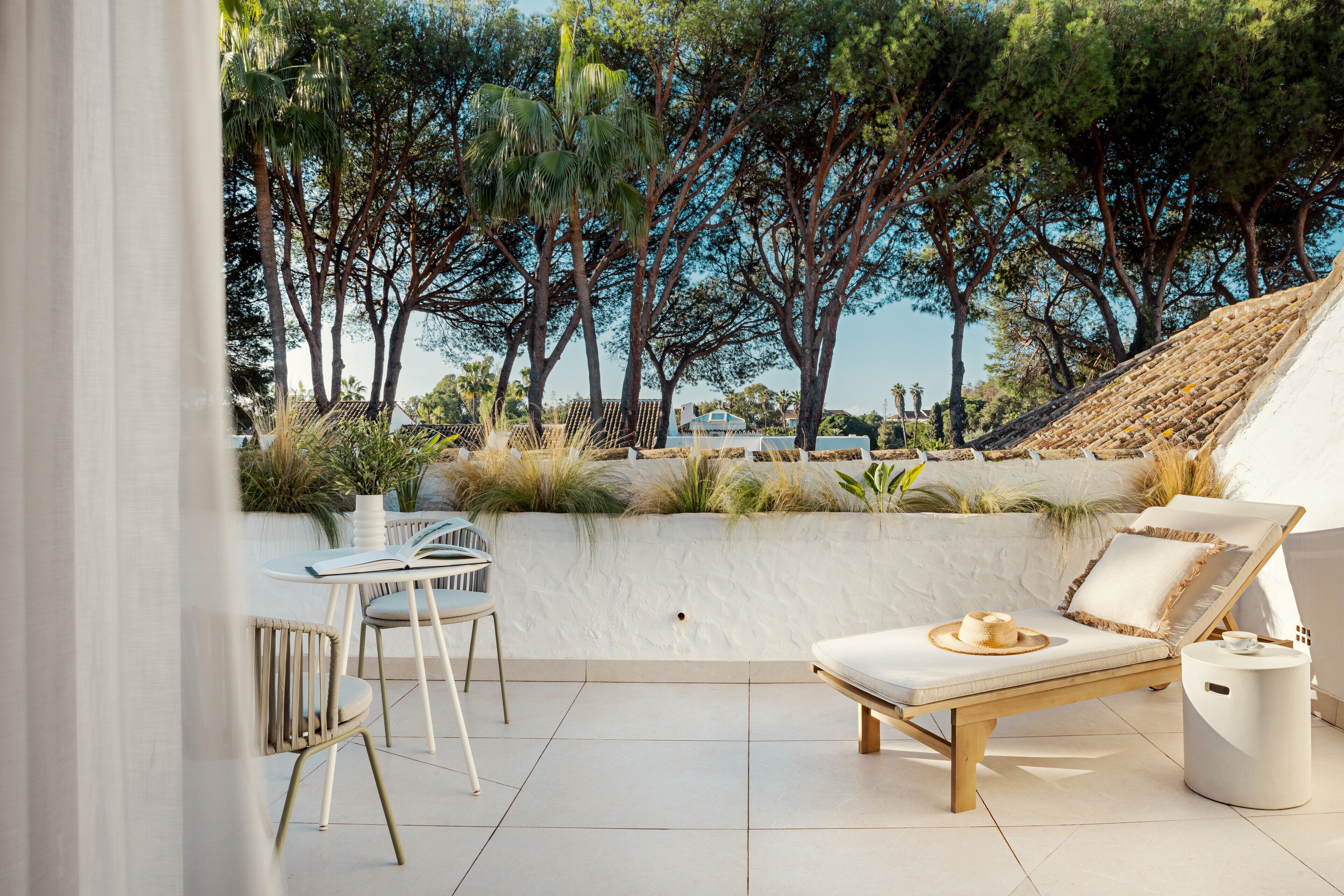 A sunlit outdoor seating area with a lounge chair, a small table, and chairs, surrounded by a white wall and lush greenery.