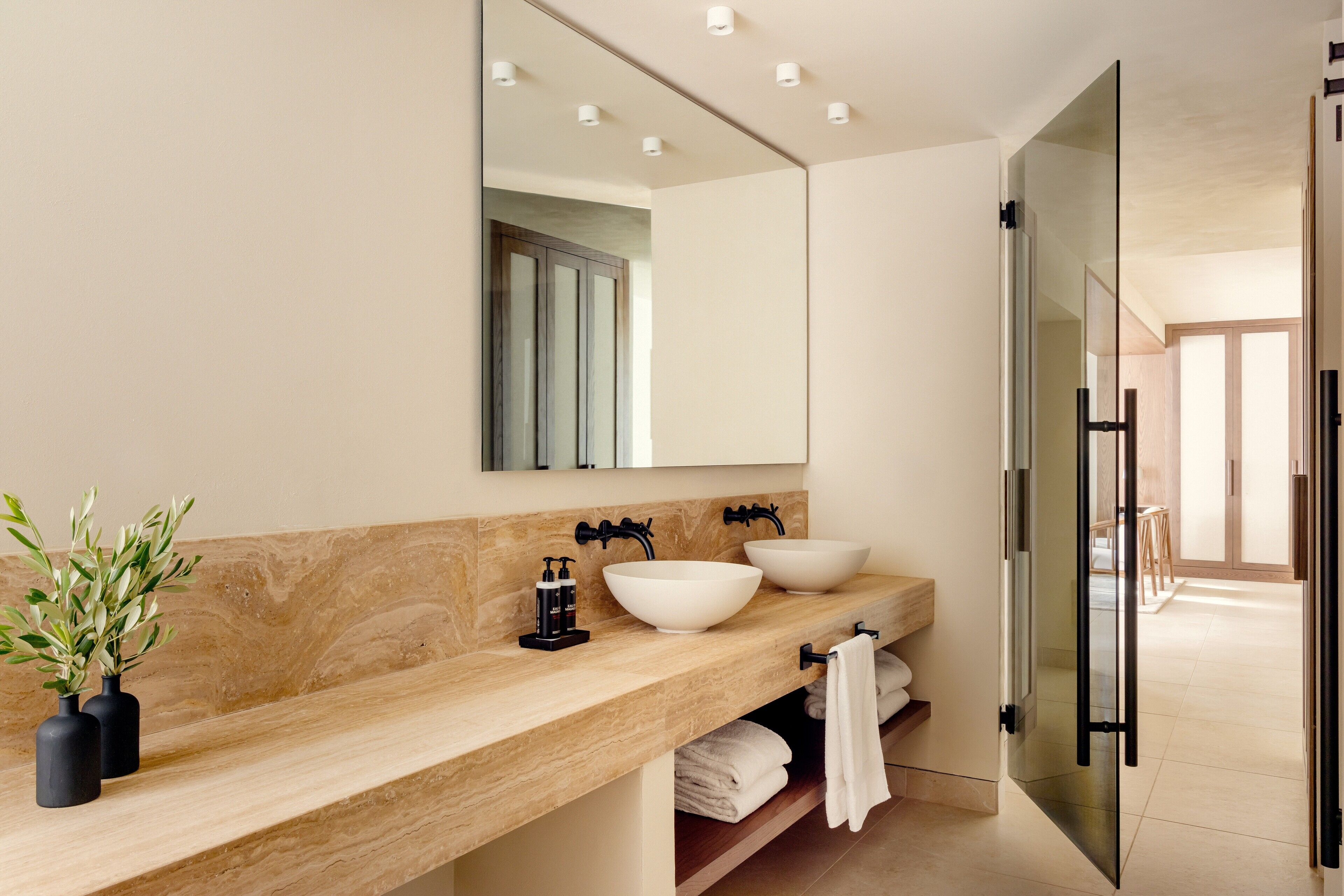 A modern bathroom with a beige marble countertop, two white vessel sinks, a large mirror, and a glass shower enclosure.