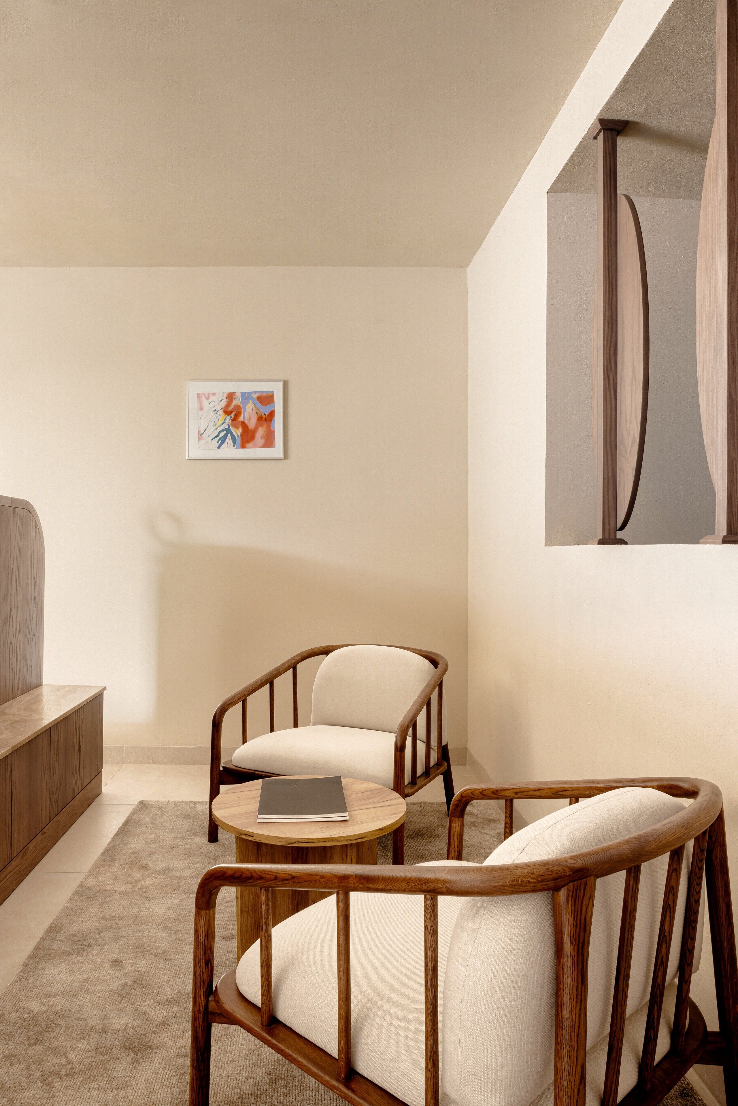 A modern living room with a wooden chair and a small round table, a framed artwork on the wall, and a large mirror.