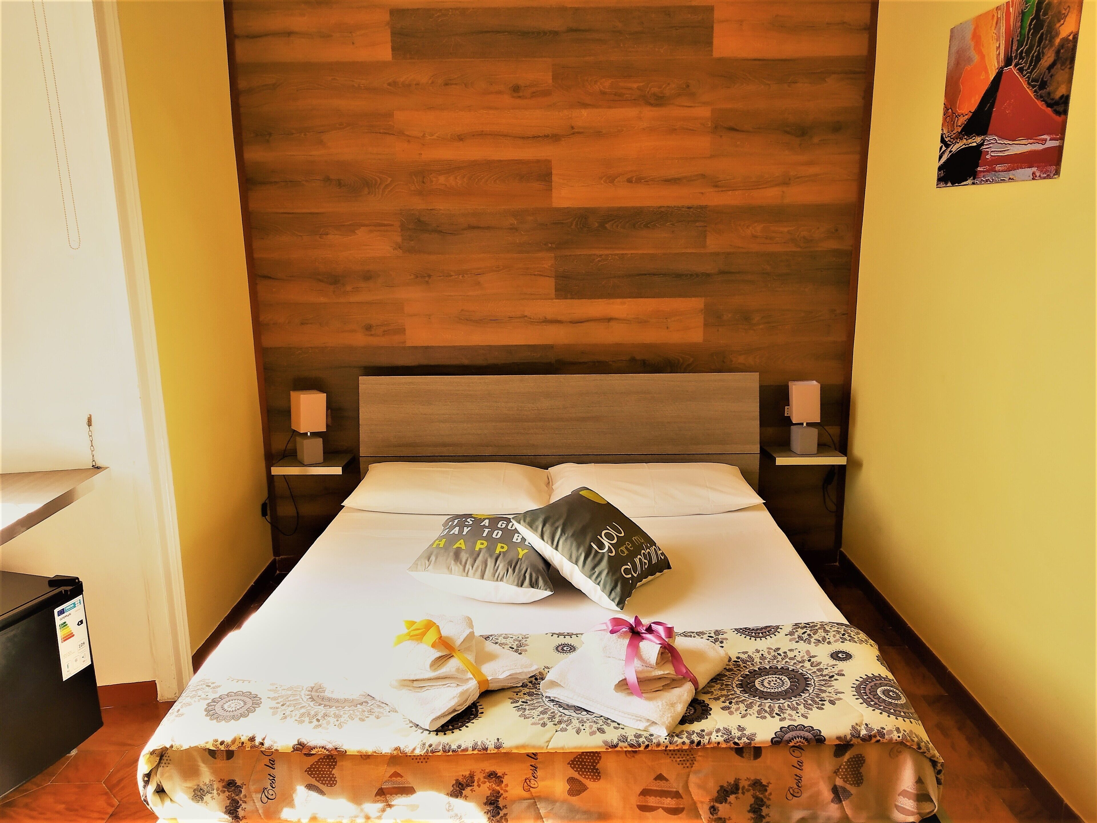 A neatly made bed with a patterned bedspread, pillows, and a decorative towel. There are two bedside tables with lamps, a wooden headboard, and a wall with a painting.