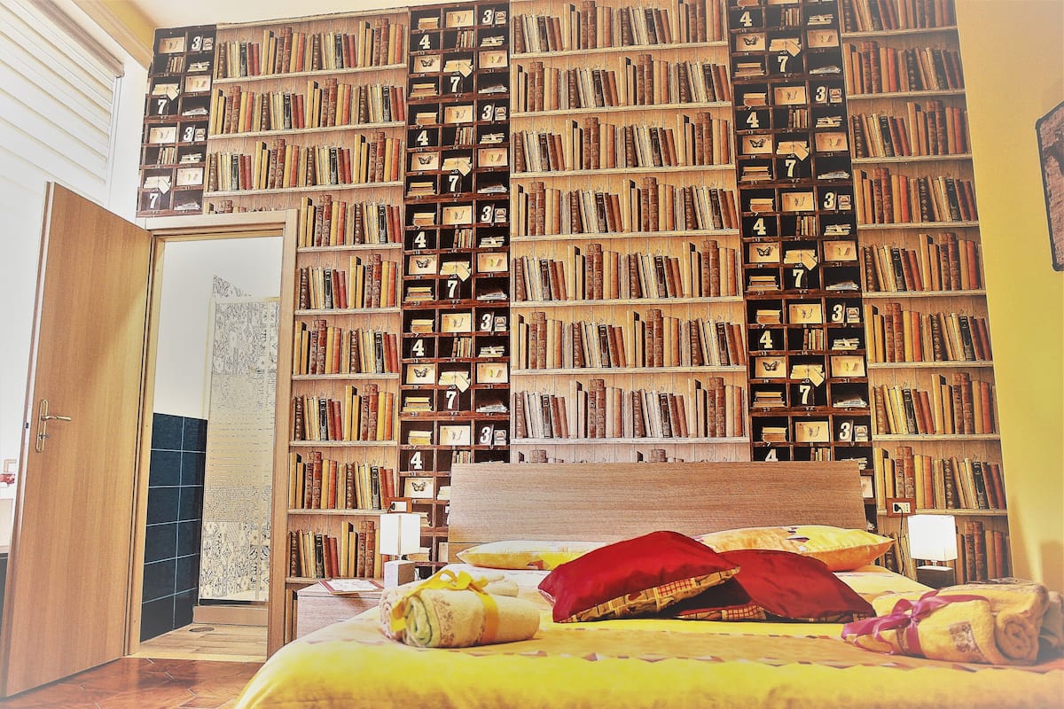 A room with a large bookshelf filled with books, a bed with yellow bedding, and a wooden door.