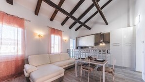 Una sala de estar acogedora con un sofá y una mesa de comedor, una cocina con armarios blancos, y un techo con vigas a la vista.