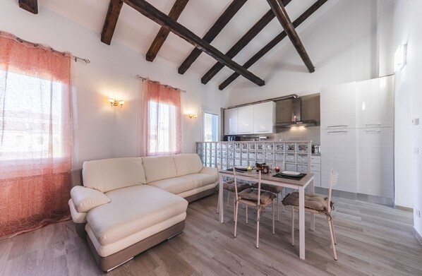 Un salon chaleureux avec un canapé et une table à manger, une cuisine dotée de meubles blancs et un plafond avec des poutres apparentes.