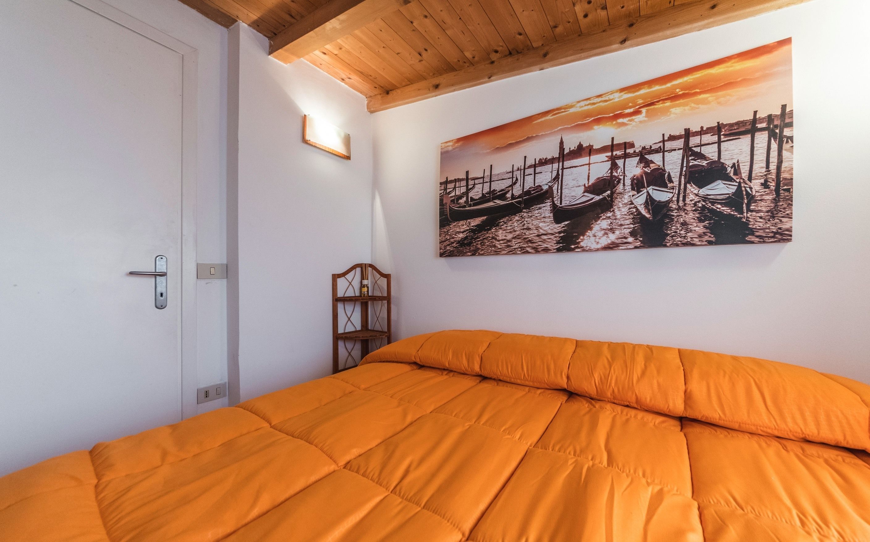 Una camera da letto con soffitto in legno, un letto con copripiumino arancione, un quadro appeso al muro raffigurante barche e una piccola sedia di legno.