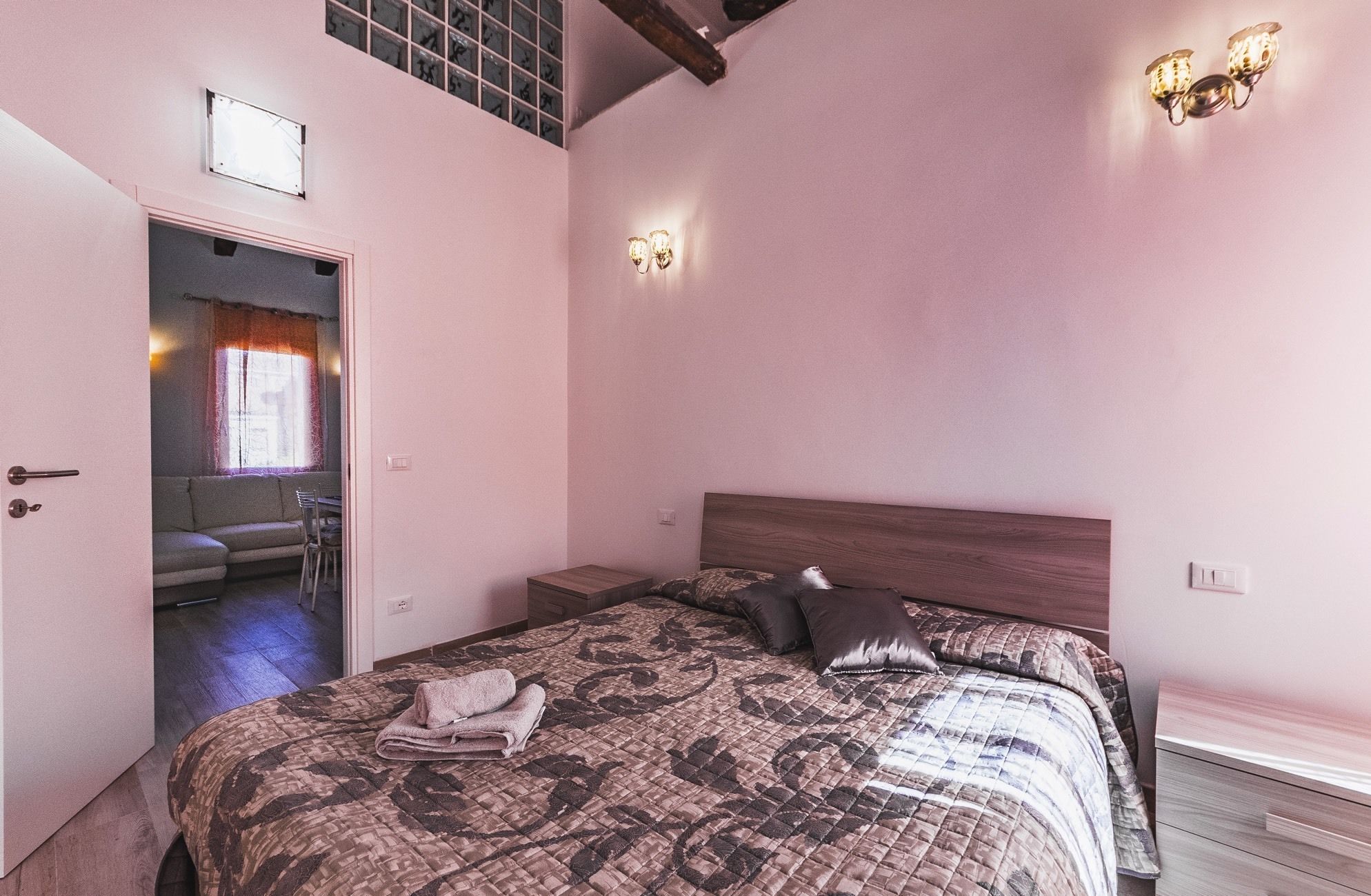 A bedroom with a bed, a nightstand, a wooden headboard, a window with curtains, and a door leading to another room.