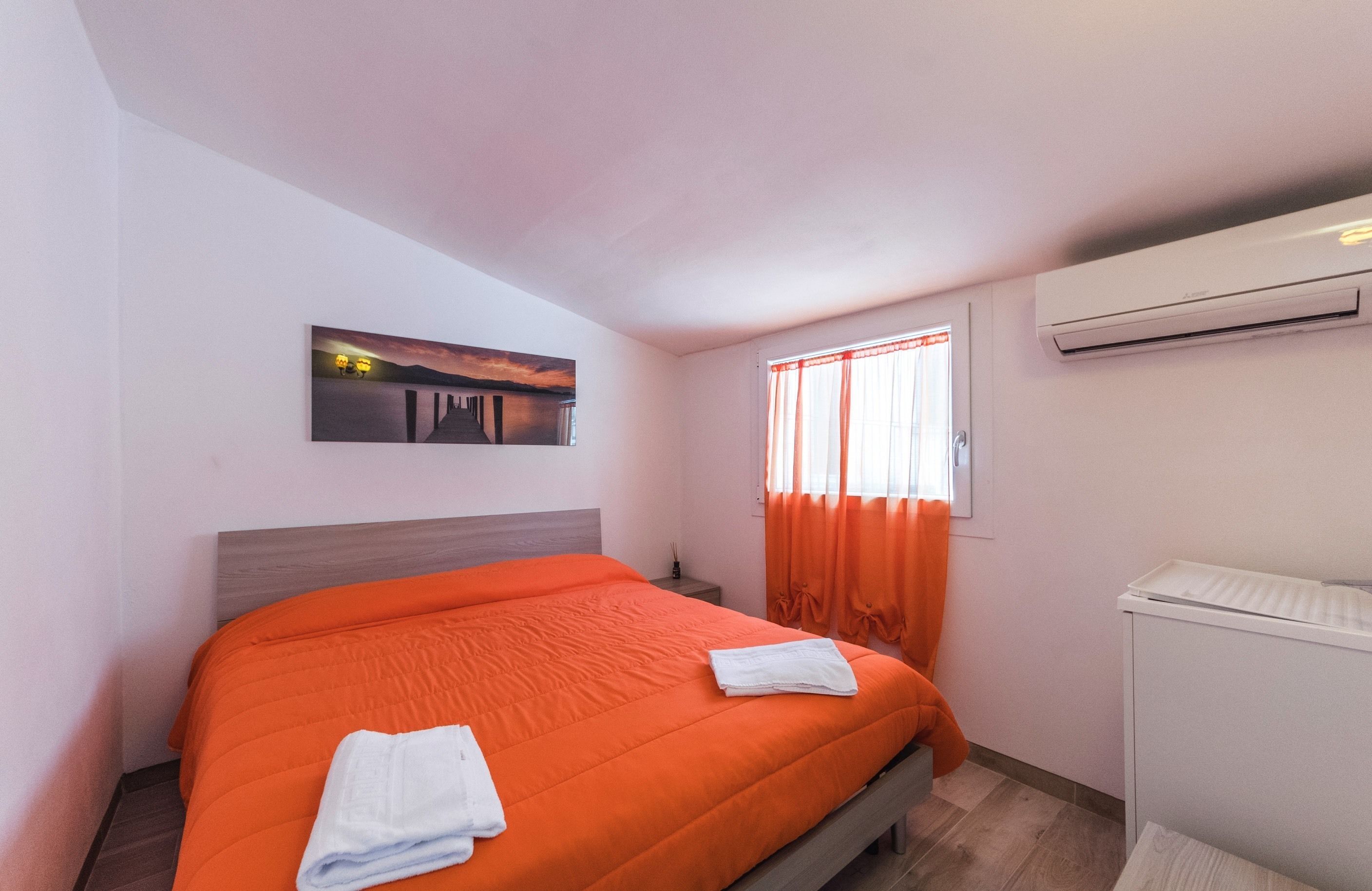 A hotel room with a bed, a window with curtains, an air conditioning unit, and a painting on the wall.