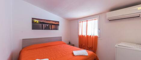 A hotel room with a bed, a window with curtains, an air conditioning unit, and a painting on the wall.