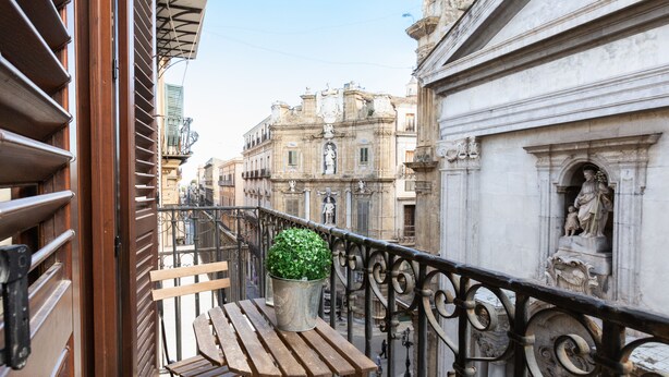 A balcony with a wooden bench and a potted plant, overlooking a street with historic buildings and a statue. - Quattro Canti Charming Flat (Palermo)