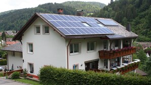Una casa de dos pisos con paneles solares en el techo, un balcón con plantas y una ladera boscosa al fondo.