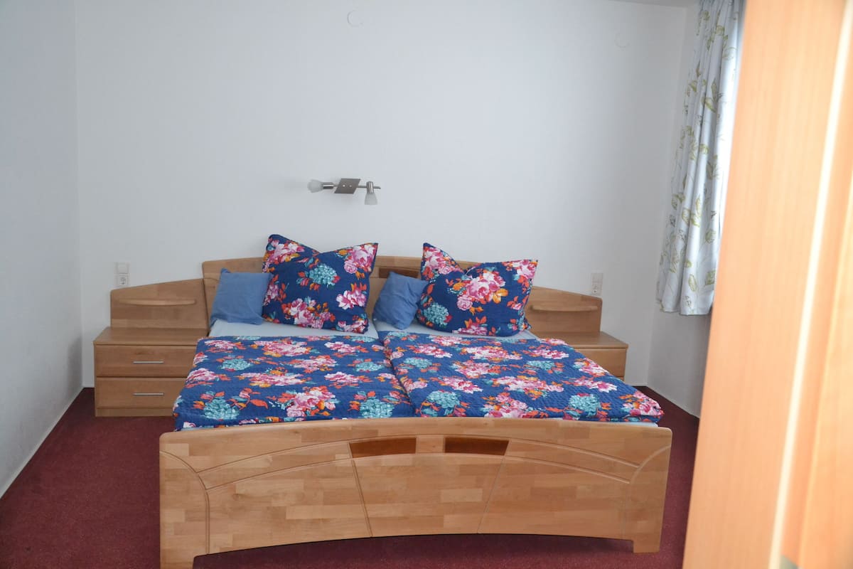 A bedroom with a wooden bed, floral bedding, a nightstand, and a wall-mounted light fixture.
