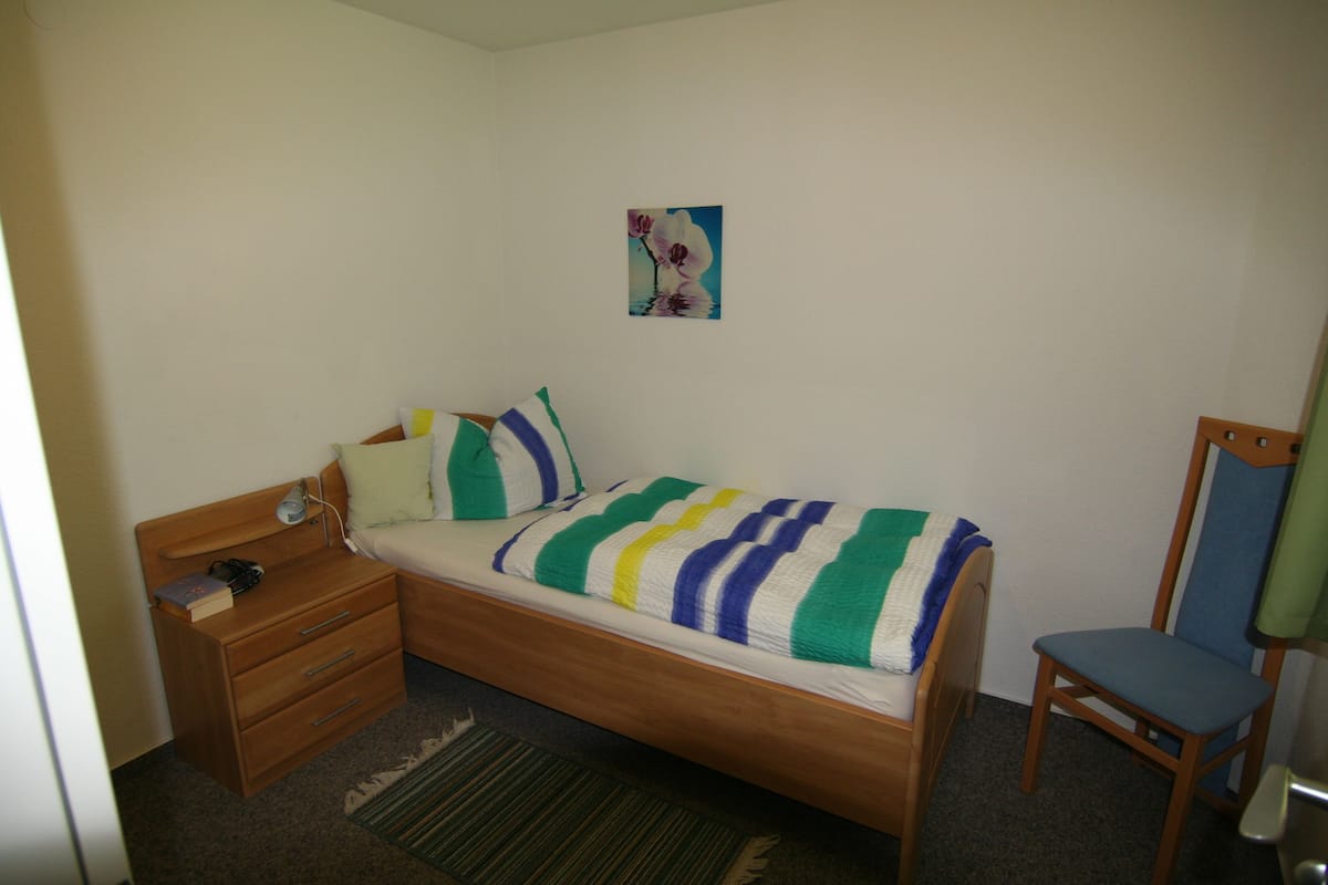 A small, neatly arranged bedroom with a single bed, a wooden nightstand, a chair, and a small table.
