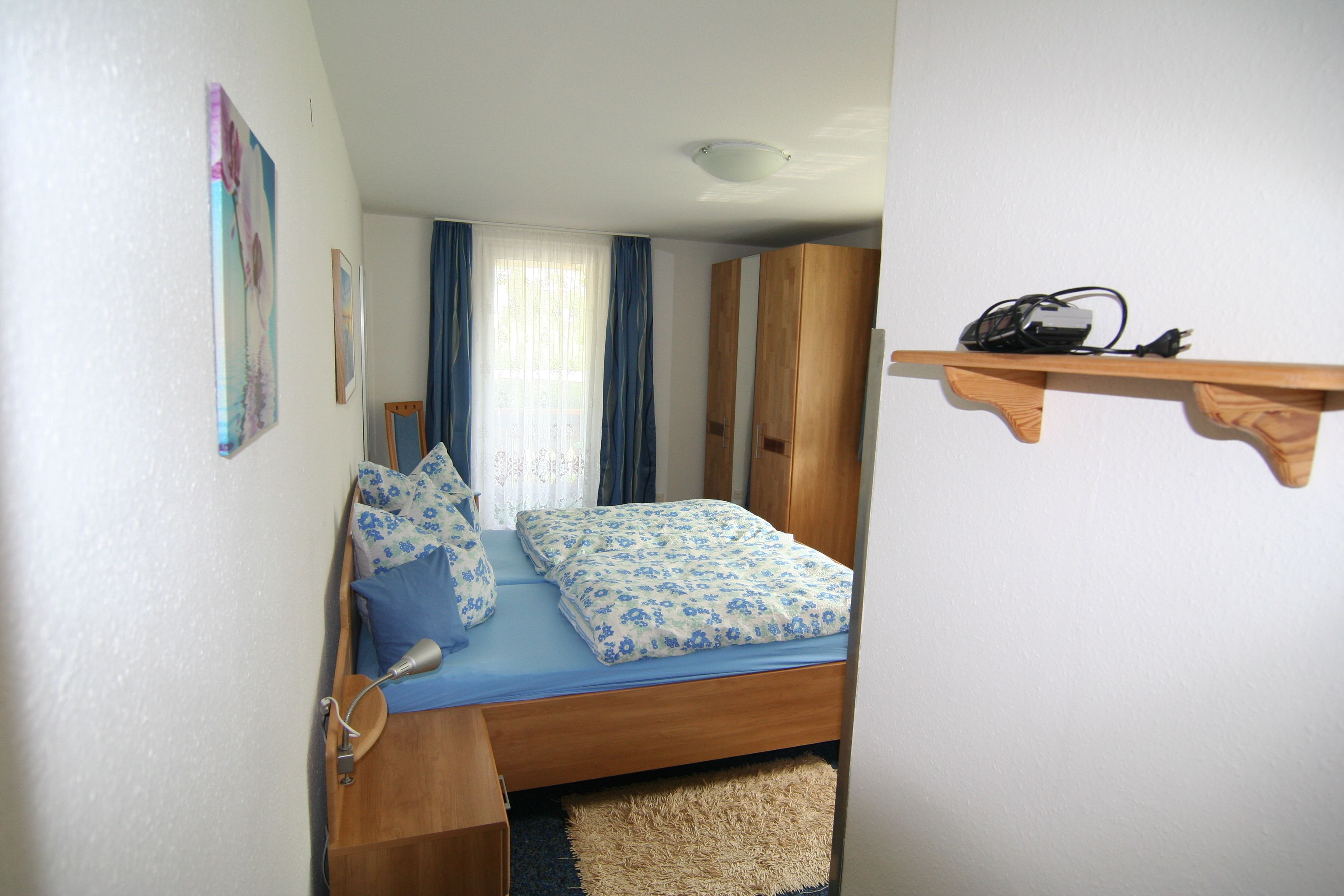 A small bedroom with a bed, a wooden wardrobe, a shelf with a power strip, and a window with curtains.