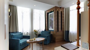 Habitación de hotel con un sofá azul, una mesa redonda de madera y un gran ventanal con cortinas.