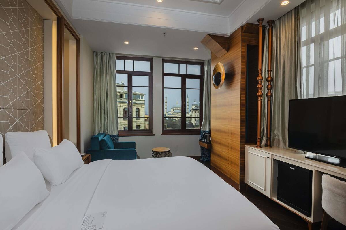 A hotel room with a large bed, a TV, a small table, and a view of city buildings.