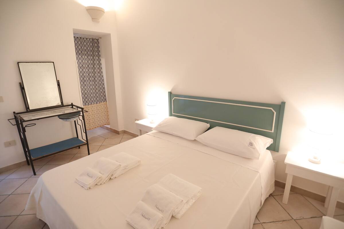 A neatly made bed with white linens and towels, a bedside table with a lamp, and a small table with a mirror and a tray.
