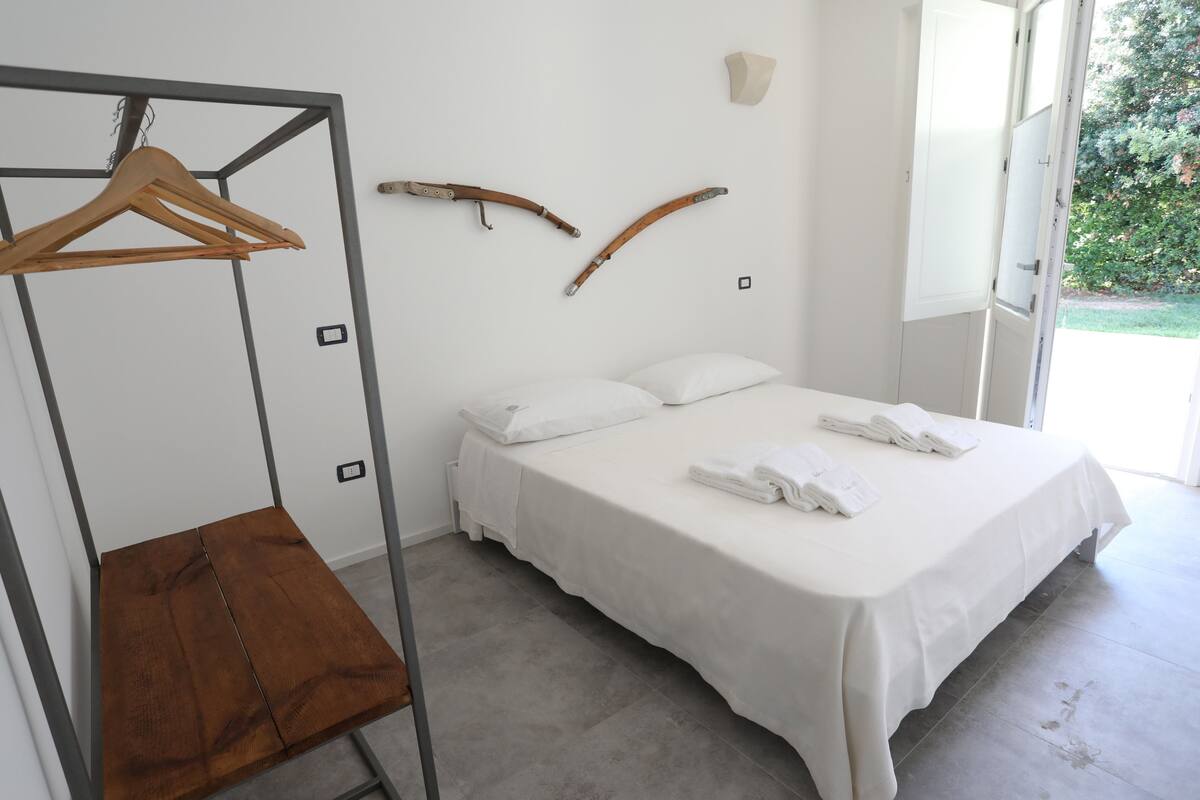 A bedroom with a bed, a wooden hanger rack, and a view of greenery outside.