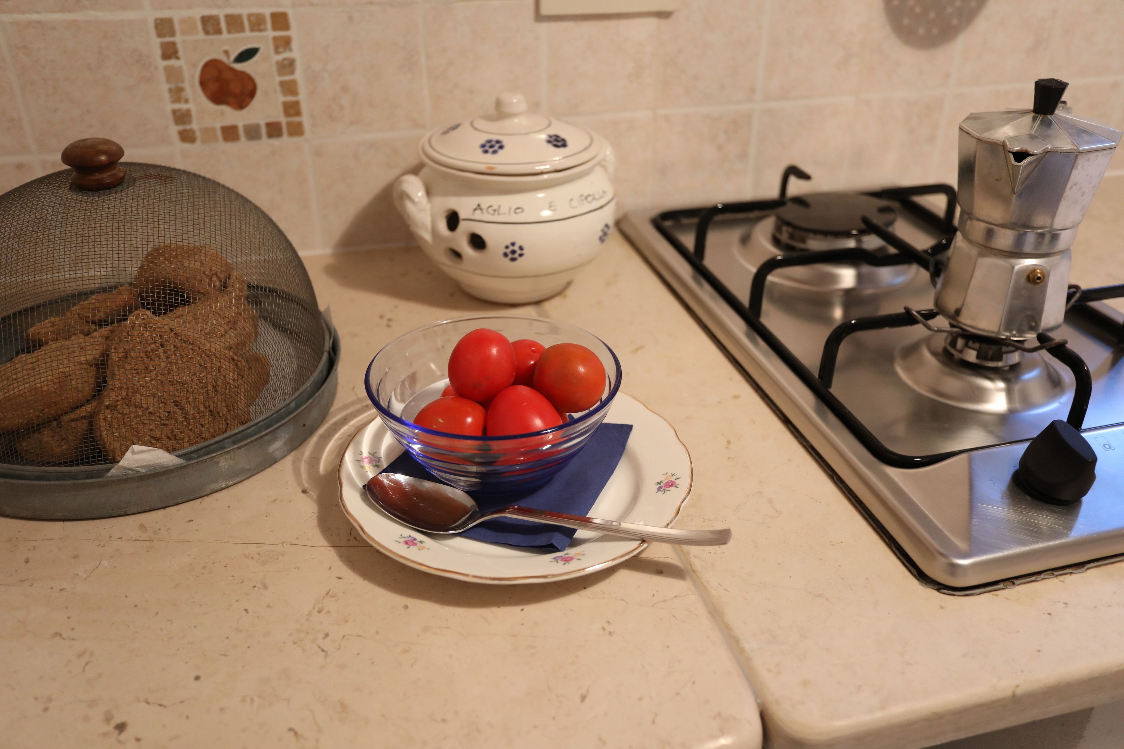 A kitchen counter with a sieve containing cookies, a bowl of tomatoes, a teapot, and a gas stove.