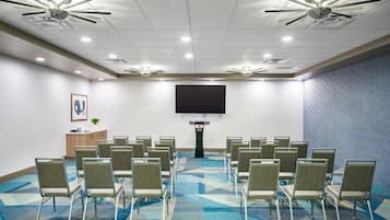 Un salón de conferencias con filas de sillas, un atril, una pantalla grande, ventiladores de techo e iluminación empotrada.