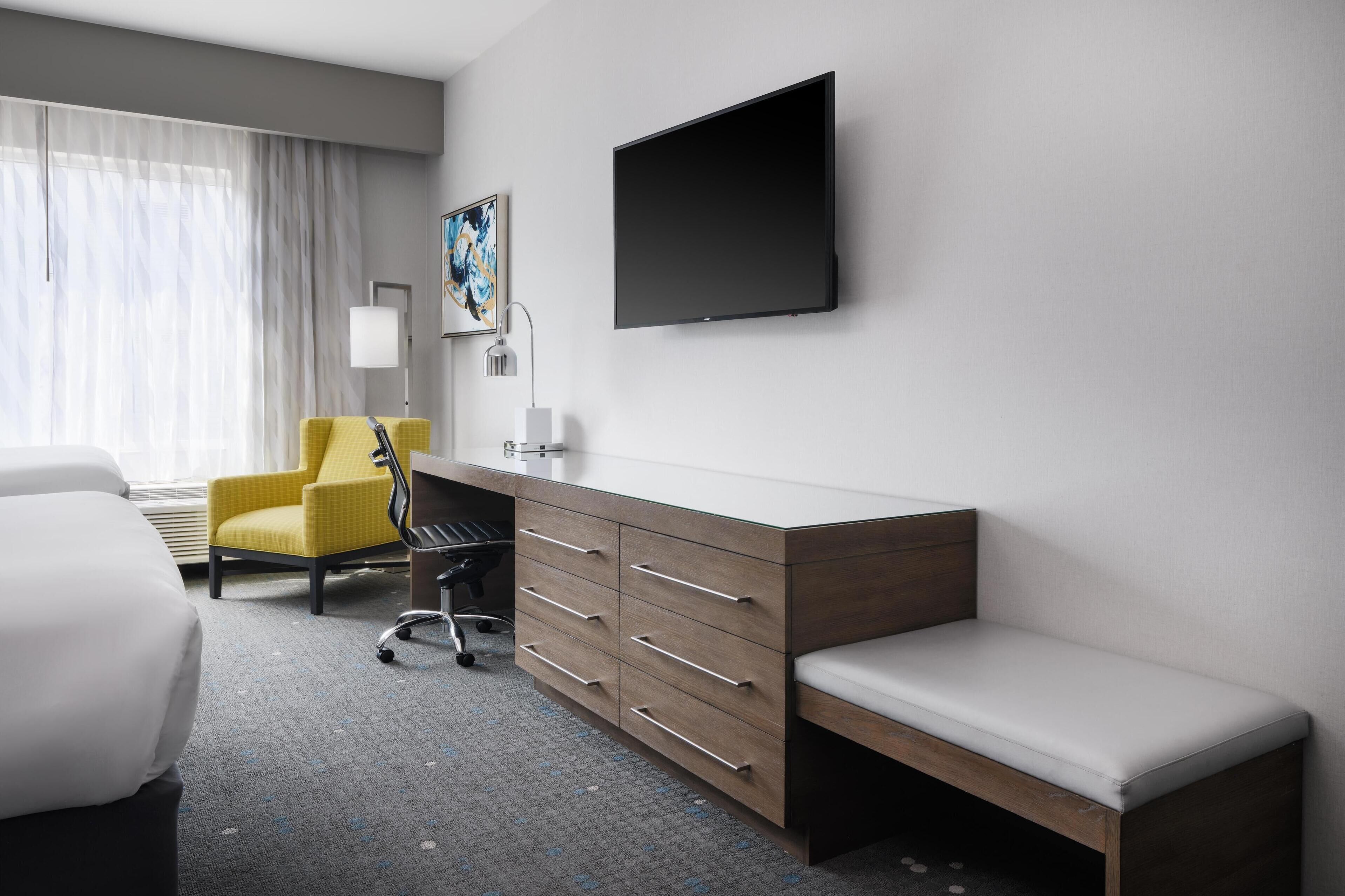 Ein Hotelzimmer mit einem Bett, einem Schreibtisch mit Schubladen, einem Sessel, einer Bank, einem Flachbildfernseher, einer Lampe und einem Gemälde an der Wand.