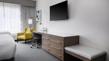 Ein Hotelzimmer mit einem Bett, einem Schreibtisch mit Schubladen, einem Sessel, einer Bank, einem Flachbildfernseher, einer Lampe und einem Gemälde an der Wand.