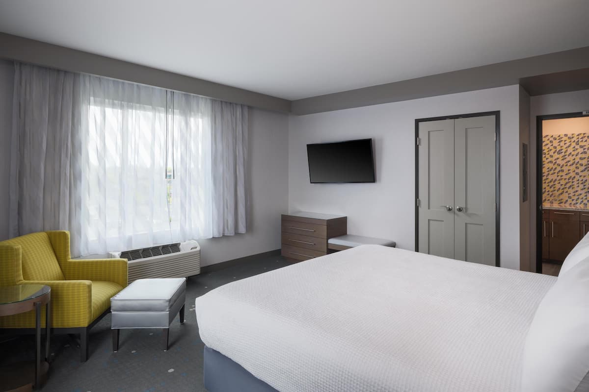 Una habitación de hotel con cama, una silla, televisión, armario y ventana con cortinas.