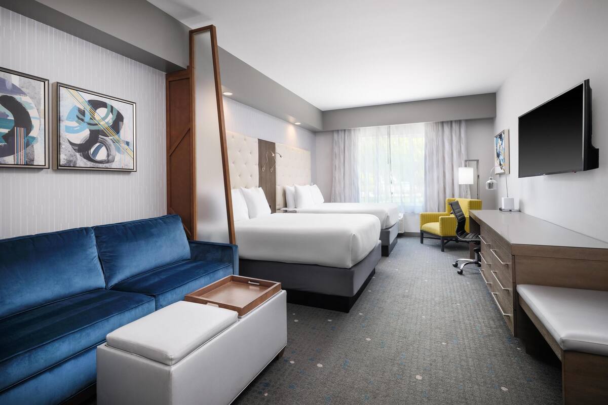 Habitación de hotel con un sofá azul, dos camas, un escritorio, una silla y obras de arte en las paredes.