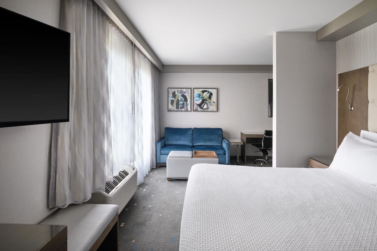 Habitación de hotel con una cama grande, un televisor fijado a la pared, un sofá azul, una mesita pequeña y dos cuadros enmarcados.
