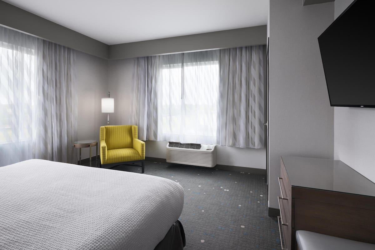 Habitación de hotel con una cama grande, un sillón amarillo, un televisor de pantalla plana y una ventana con cortinas.