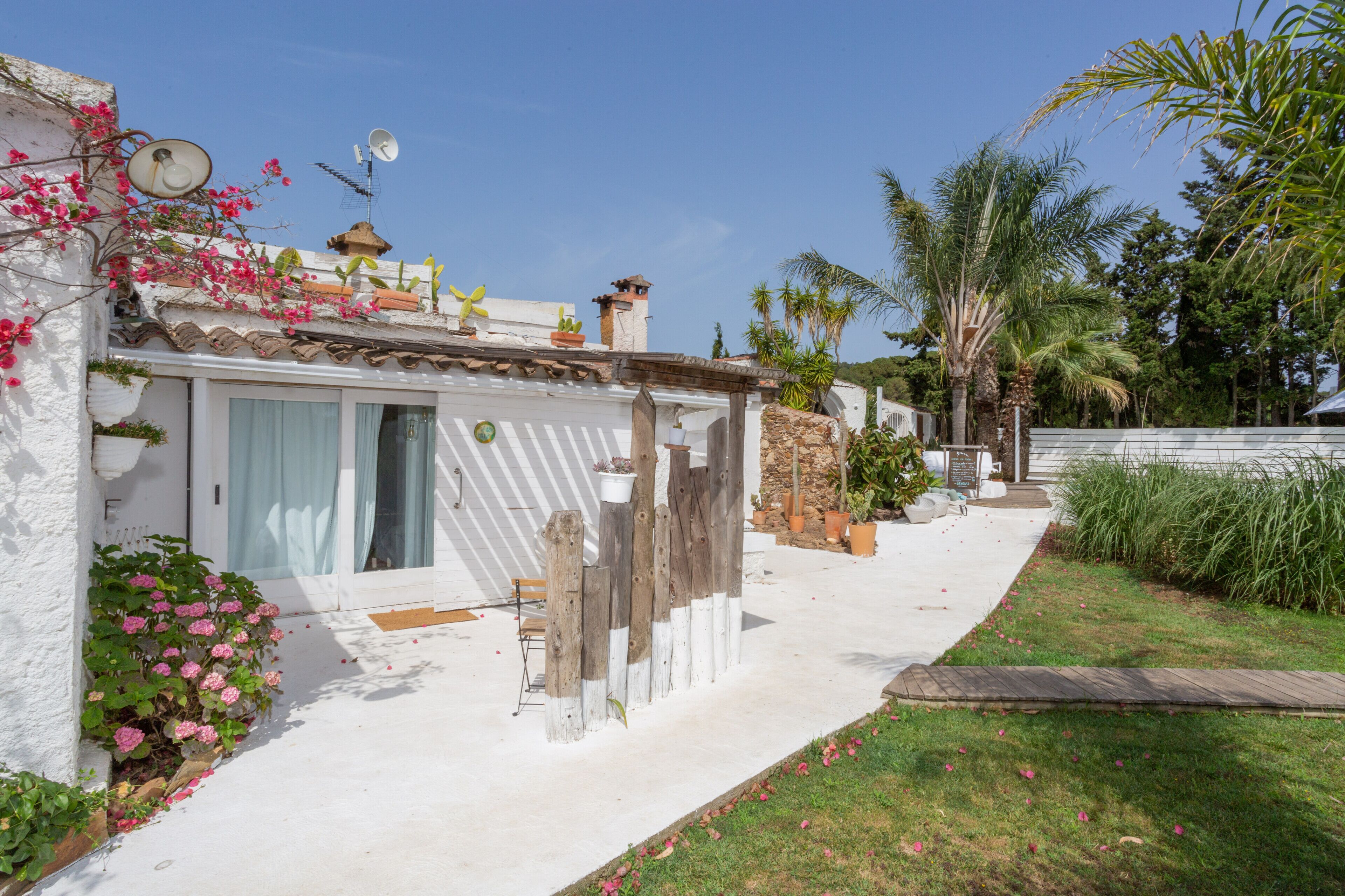 Une maison à l’extérieur blanc, avec une clôture en bois et un jardin agrémenté de fleurs roses et de palmiers.