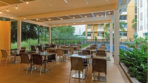 An outdoor cafe area with tables and chairs, a covered seating area, and a view of a building and greenery.