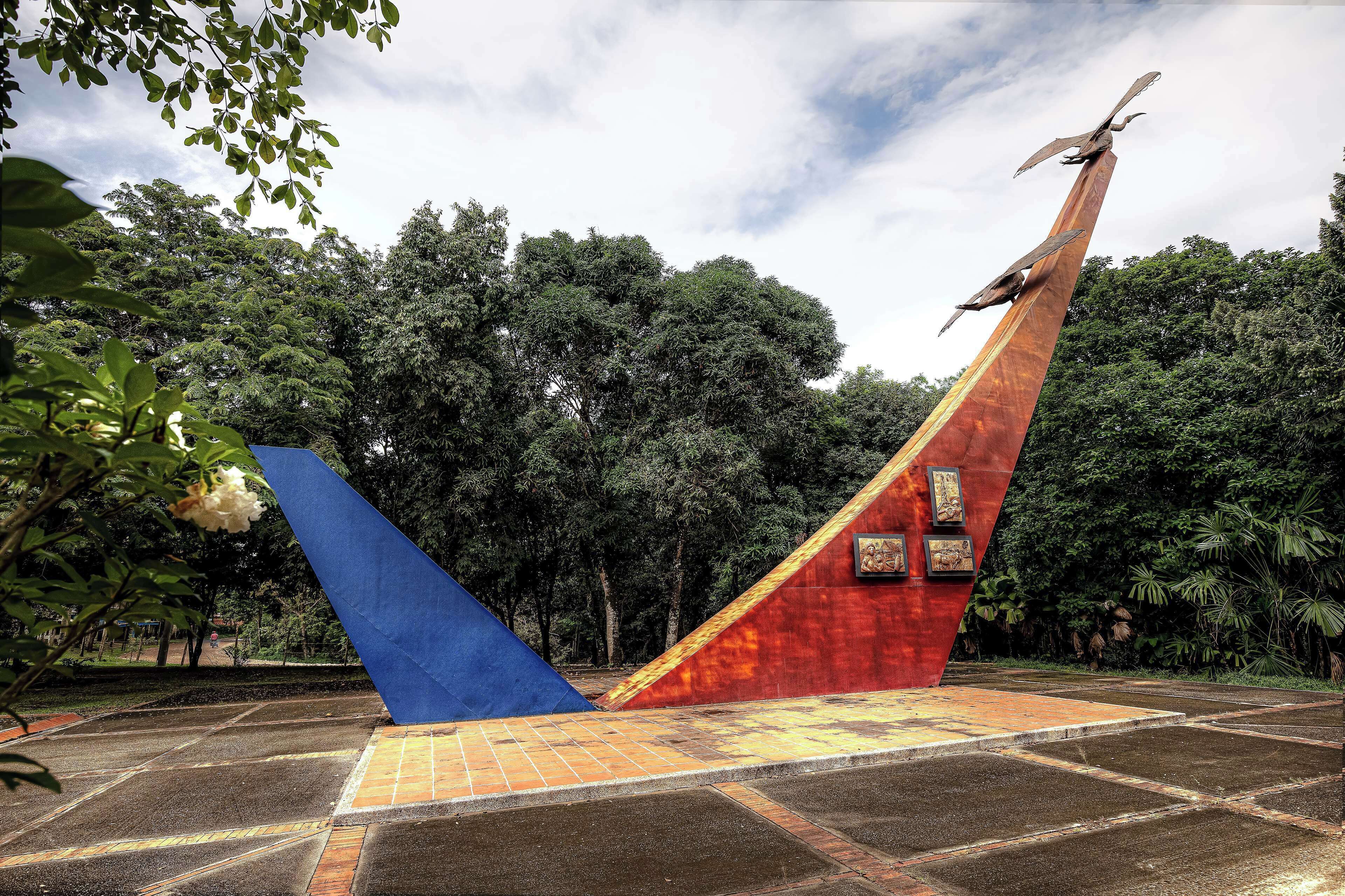 A sculpture of birds on a tall red and blue structure in a park with trees in the background.