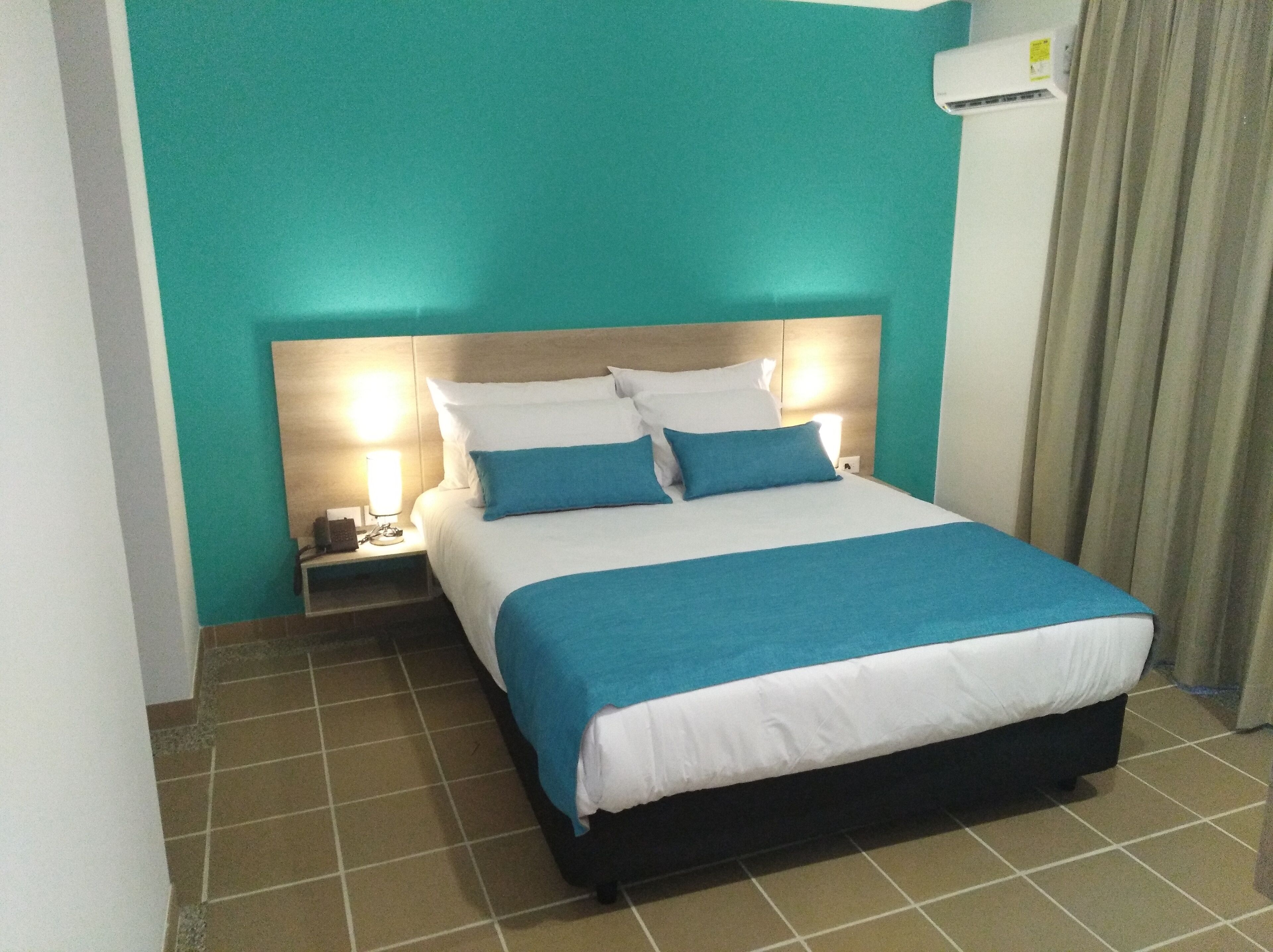 A hotel room with a bed, a nightstand, a lamp, and an air conditioning unit.