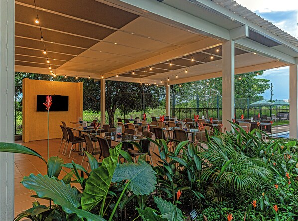 Un patio al aire libre con mesas y sillas, rodeado de abundante vegetación y vistas al cielo.