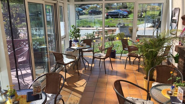 A sunlit patio area with wicker chairs and tables, large windows offering views of parked cars and a garden.