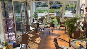 A sunlit patio area with wicker chairs and tables, large windows offering views of parked cars and a garden. - Café & Pension Meine Sonne ... Sole Mio (Bad Sooden-Allendorf)