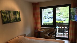 A hotel room with a bed, a small table with a chair, a window with a view of parked cars and greenery outside, and a painting on the wall.